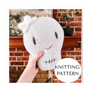 May include: A light grey, knitted skull-shaped plush toy with black embroidered eyes, a stitched mouth, and a white bow. The text "KNITTING PATTERN" is in a white circle.