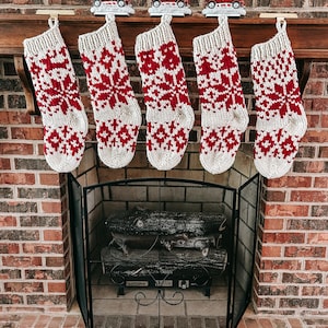 Puede incluir: Cinco calcetines navideños de punto con diseños rojos y blancos cuelgan sobre una chimenea. Camiones rojos decorativos con árboles de Navidad están sobre la repisa. Los calcetines tienen un diseño de copo de nieve y geométrico.