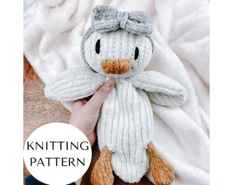 May include: White knitted plush toy duck with a gray bow, brown feet, and black eyes. The duck is sitting on a white fuzzy blanket.