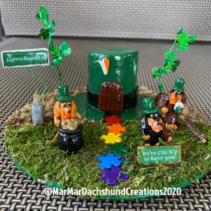 May include: A whimsical St. Patrick's Day scene featuring three dachshund figurines dressed as leprechauns. The scene includes a green top hat, a pot of gold, a rainbow path, and a sign that reads "We're lucky to have you!"