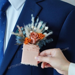 Puede incluir: Una chaqueta de traje azul con una camisa blanca y una corbata azul con estampado. Una mano sostiene un pequeño ramo de flores secas con un respaldo de tela melocotón. El ramo se está prendiendo al ojal de la chaqueta.