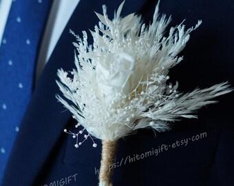 May include: A white dried flower boutonniere with a single white rose and pampas grass, tied with twine. The boutonniere is pinned to a dark blue suit jacket.