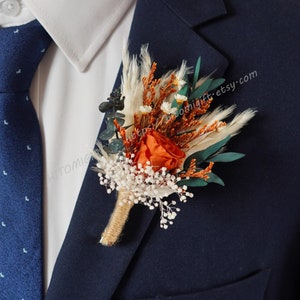 May include: A boutonniere with dried flowers in shades of orange, white, and brown. The boutonniere includes a single orange rose, white baby's breath, dried grasses, and eucalyptus leaves. The boutonniere is pinned to a navy blue suit jacket.