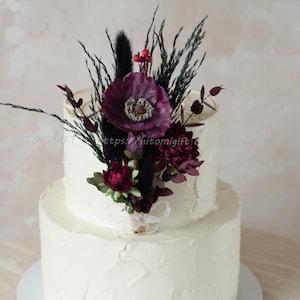 Könnte beinhalten: Zweistöckiger weißer Kuchen mit Buttercreme-Frosting, verziert mit einem Strauß burgunderroter und schwarzer Blumen. Der Strauß enthält burgunderrote Anemonen, burgunderrote Rosen, schwarze getrocknete Gräser und schwarzes Pampasgras.
