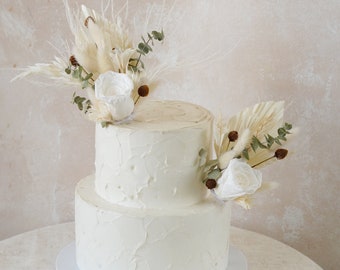 May include: Two-tiered white wedding cake with buttercream frosting and a rustic floral arrangement of dried flowers and greenery.