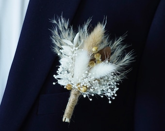 May include: A small bouquet of dried flowers in shades of white and brown, with a touch of gold, is pinned to a dark blue suit jacket.
