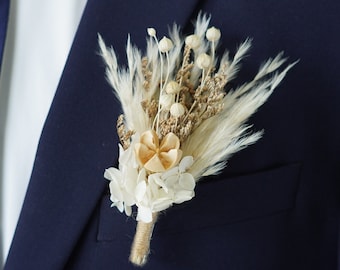 May include: A white and beige dried flower boutonniere with pampas grass, perfect for a wedding or special occasion.