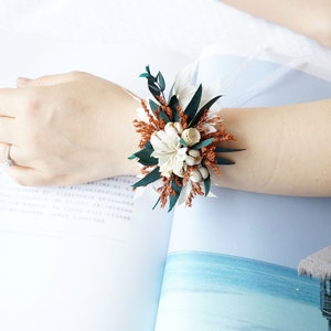 May include: A wrist corsage made with dried flowers. The corsage is made with white flowers, brown dried grasses, and green leaves.