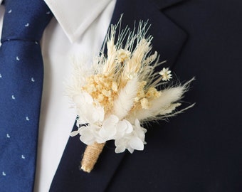 May include: A white and cream dried flower boutonniere with a burlap wrap. The boutonniere is pinned to a navy blue suit jacket.