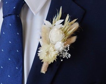 May include: A white and beige dried flower boutonniere with a navy blue suit jacket.