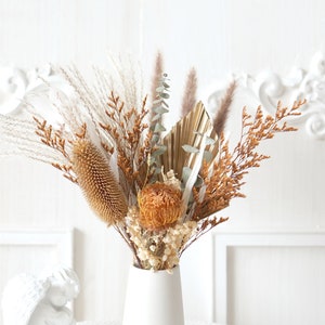 Può includere: Un arrangiamento di fiori secchi in un vaso bianco. Il bouquet comprende erbe secche, eucalipto e una varietà di fiori secchi in tonalità di marrone e arancio.