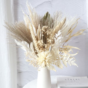 Può includere: Un vaso di ceramica bianca riempito con un bouquet di erba della pampa secca, code di coniglio e altri fiori secchi. Il vaso è appoggiato su un tavolo bianco con una rivista sopra.