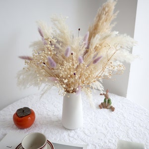 Pu&ograve; includere: Un vaso di ceramica bianco contiene una composizione floreale secca. La composizione comprende soffice erba della pampa beige, delicato velo da sposa bianco e morbida erba coda di coniglio lavanda. Un frutto arancione e una statuina di coniglio in ceramica sono sul tavolo.