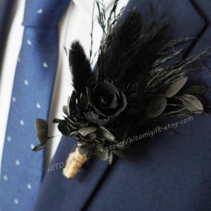 以下が含まれることがあります： 結婚式や特別な日に最適な、黒の羽根とバラのブートニアに緑の葉が添えられています。