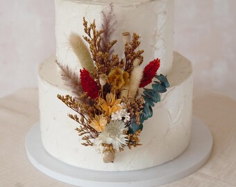 May include: Two-tiered white cake with textured frosting, decorated with a bouquet of dried flowers. The arrangement includes red, yellow, and cream-colored blooms, along with green eucalyptus leaves and tan grasses. The cake sits on a white cake stand.