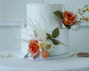 May include: A two-tiered white cake with textured frosting, adorned with orange and white roses, green leaves, and feathery accents. The cake sits on a white cake stand, perfect for a wedding or celebration.