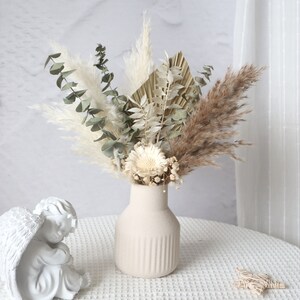 Può includere: Un arrangiamento di fiori secchi in un vaso di ceramica bianco con una superficie testurizzata. L'arrangiamento include erba della pampa secca, eucalipto e fiori bianchi.