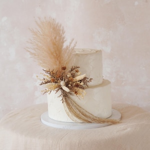 Pode incluir: Um bolo branco de dois andares decorado com flores secas e capim-dos-pampas. O bolo está em um suporte branco e é exibido em uma toalha de mesa bege. O bolo é de cor clara e tem uma superfície texturizada.