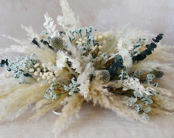 May include: A dried floral arrangement featuring pampas grass, white flowers, and accents of blue-green foliage. The arrangement is a mix of textures and colors, creating a natural and elegant aesthetic. Perfect for home decor or special events.