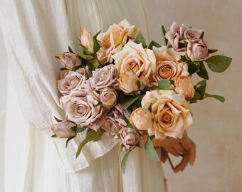 May include: A bouquet of artificial roses in shades of peach, mauve, and cream. The roses are in various stages of bloom, from tightly closed buds to fully open flowers. Green leaves provide contrast. The bouquet is held against a white background.
