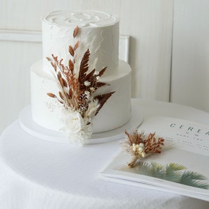 May include: A two-tiered white wedding cake decorated with dried flowers and greenery. The cake is on a white cake stand and is sitting on a white tablecloth. There is a magazine on the table with the word "Cereal" printed on the cover.