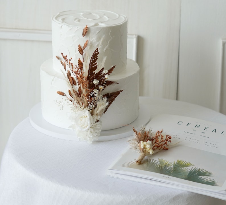 May include: A two-tiered white wedding cake decorated with dried flowers and greenery. The cake is on a white cake stand and is sitting on a white tablecloth. There is a magazine on the table with the word "Cereal" printed on the cover.