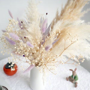 Pu&ograve; includere: Un vaso bianco pieno di una composizione floreale secca. La composizione include soffice erba della pampa beige, delicato velo da sposa bianco e morbida erba coda di coniglio viola. Un piccolo frutto arancione e un coniglio di ceramica sono sul tavolo.