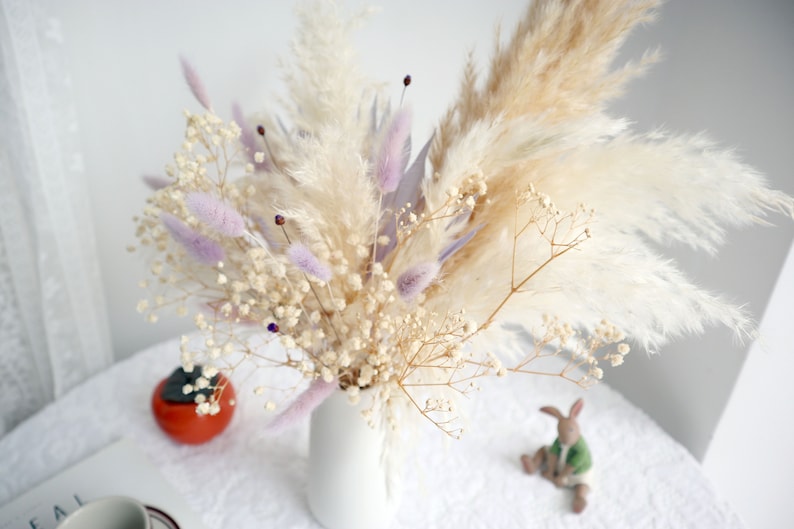 Pu&ograve; includere: Un vaso bianco pieno di una composizione floreale secca. La composizione include soffice erba della pampa beige, delicato velo da sposa bianco e morbida erba coda di coniglio viola. Un piccolo frutto arancione e un coniglio di ceramica sono sul tavolo.