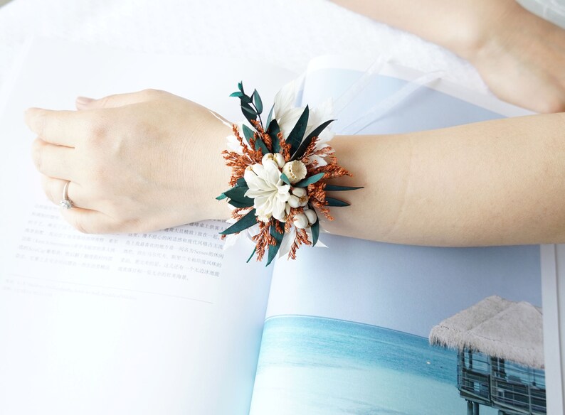 May include: A wrist corsage made with dried flowers. The corsage is made with white flowers, brown dried grasses, and green leaves.