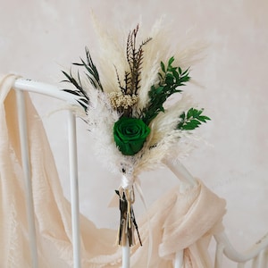 Op de afbeelding: Een boeket van gedroogde bloemen met een levendige groene roos als middelpunt. Het arrangement bevat pluizig wit pampasgras, donkergroene bladeren en bruine accenten, vastgebonden met een lint. Het boeket rust op een witte metalen stoel.