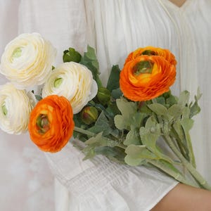 May include: A bunch of artificial ranunculus flowers. The bouquet features white and orange blooms with green leaves. The flowers are held against a white background.