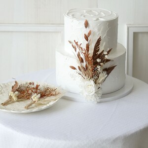 May include: A two-tiered white wedding cake with a rustic, dried floral arrangement on top. The cake is on a white cake stand and the flowers are a mix of brown and white.
