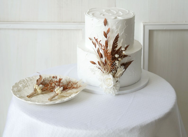 May include: A two-tiered white wedding cake with a rustic, dried floral arrangement on top. The cake is on a white cake stand and the flowers are a mix of brown and white.