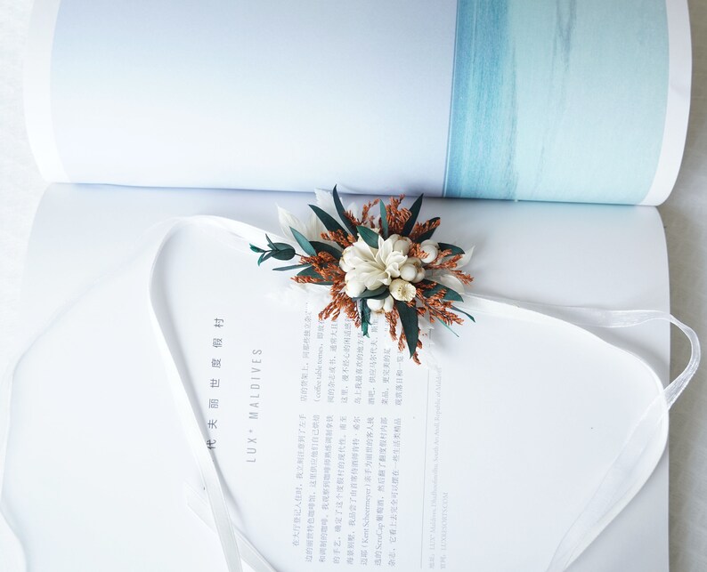 May include: A white and brown dried flower arrangement with green leaves, tied with a white ribbon. The arrangement is on a white book with the text "LUX MALDIVES" printed on the cover.