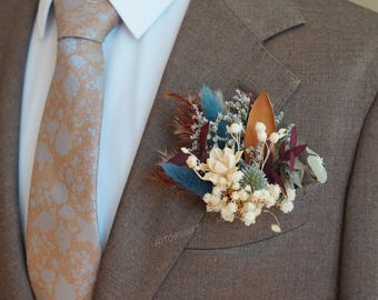 Puede incluir: Chaqueta de traje marrón con corbata estampada y un prendido floral. El prendido presenta plumas azules, flores blancas y follaje seco. La corbata tiene un estampado marrón claro y blanco.