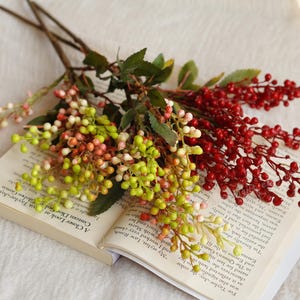 Może przedstawiać: Bukiet sztucznych jagód w odcieniach czerwieni, zieleni i różu spoczywa na otwartej książce. Jagody są zgrupowane na brązowych łodygach z zielonymi liśćmi. Strony książki wyświetlają drukowany tekst.