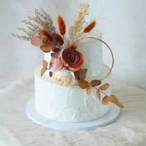 May include: Two-tiered white wedding cake decorated with dried flowers and greenery. The cake is topped with a gold ring and a cluster of dried flowers, including a large orange rose.