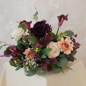 May include: A vibrant floral display with a deep burgundy rose, cream roses, peach dahlias, and dark purple calla lilies. The arrangement includes green foliage and small white flowers, creating a full, textured bouquet.