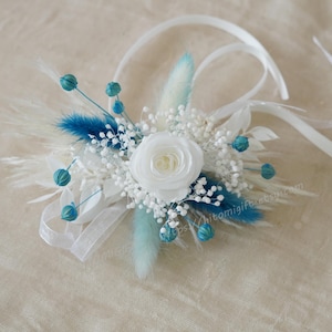 May include: A white silk flower with blue and white dried flowers and white ribbon. The bouquet is tied with a white ribbon.