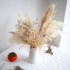 Pu&ograve; includere: Un vaso di ceramica bianco pieno di una composizione floreale secca. La composizione include soffice erba della pampa beige, delicato velo da sposa bianco e morbidi accenti viola. Il vaso &egrave; su una tovaglia di pizzo bianco.