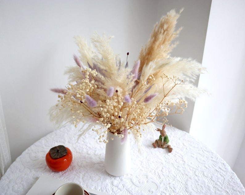 Pu&ograve; includere: Un vaso di ceramica bianco pieno di una composizione floreale secca. La composizione include soffice erba della pampa beige, delicato velo da sposa bianco e morbidi accenti viola. Il vaso &egrave; su una tovaglia di pizzo bianco.