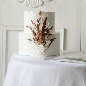 May include: A two-tiered white wedding cake decorated with dried flowers and a single white rose.