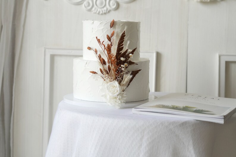 May include: A two-tiered white wedding cake decorated with dried flowers and a single white rose.