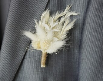May include: A white dried flower boutonniere with a brown twine wrap. The boutonniere is made of dried pampas grass and other dried flowers.