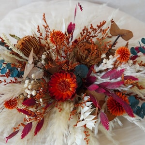 Könnte beinhalten: Eine Trockenblumen-Arrangement mit weißem Pampasgras, burgunderroten und orangefarbenen Trockenblumen und blauen Eukalyptusblättern.