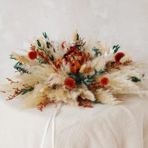 Peut inclure: Une composition florale séchée avec une grande fleur orange au centre, entourée de pampas blancs et de petites fleurs orange. L'arrangement est posé sur une nappe en lin blanc.