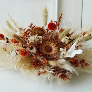 Puede incluir: Un arreglo de flores secas con una mezcla de flores y hierbas secas blancas, marrones y naranjas. El arreglo tiene forma redonda y es perfecto para una pieza central.