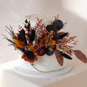 May include: A white frosted cake adorned with a colourful arrangement of dried flowers and foliage. The arrangement features orange, black, and burgundy elements, with a gold ring at the base.