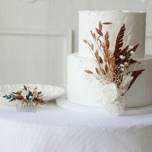 May include: Two-tiered white wedding cake decorated with dried flowers and greenery. The cake is on a white cake stand and a white tablecloth. A small floral hair comb is on a white plate to the left of the cake.