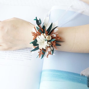 May include: A wrist corsage made with dried flowers and greenery. The corsage features white flowers, brown dried grasses, and green leaves.